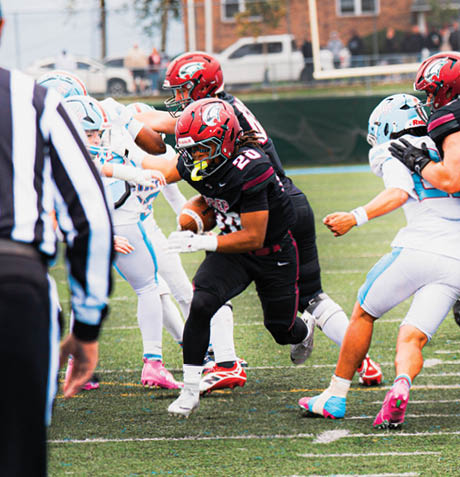 A group of football players in red and black uniforms and white and blue uniforms are actively engaged in a play on a green field with a referee partially visible in the foreground. Description generated by AI