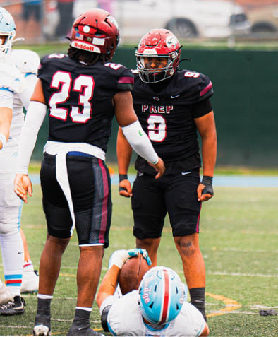 Two people in black football uniforms stand on a field while a person in a light blue helmet lies on the ground holding a football. Description generated by AI