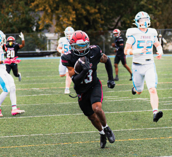 An adult in a black football uniform with a red helmet is running with the ball on a green field while several adults in white and black uniforms chase behind. Description generated by AI