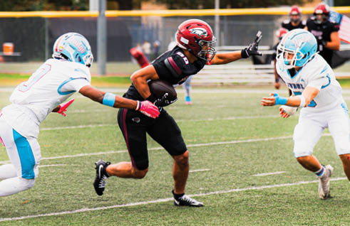 A person in a black football uniform is running with the ball while two people in white and blue football uniforms attempt to block or tackle them on a grassy field. Description generated by AI
