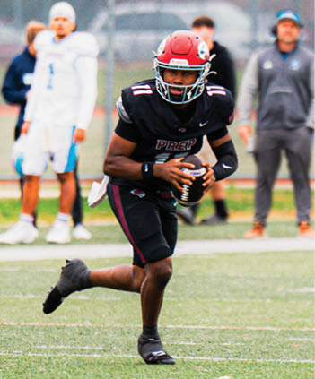 A person in a black football uniform and red helmet is running on a field while holding a football, with other people blurred in the background. Description generated by AI