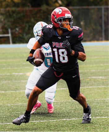 A person in a black football uniform with the number 18 is holding a football and appears to be running or preparing to throw while another person in a white uniform is behind them on a football field. Description generated by AI