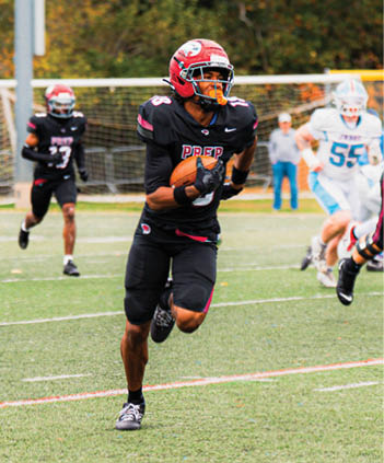 A person in a black football uniform with a red helmet is running on a field while holding a football, with other players and a goalpost visible in the background. Description generated by AI