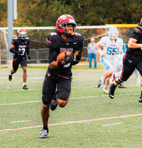 A person in a black football uniform with a red helmet is running on a field while holding a football, with other players and a goalpost visible in the background. Description generated by AI