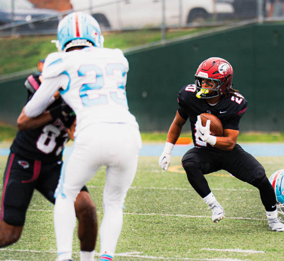 A person in a black football uniform with a red helmet is holding a football and preparing to run while two other players in white and black uniforms are nearby on a grassy field. Description generated by AI