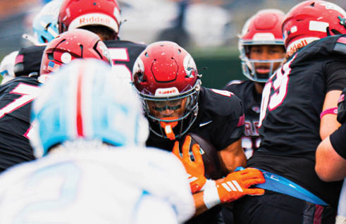 People wearing football helmets and uniforms are engaged in a play with one person holding the ball tightly while others surround them. Description generated by AI