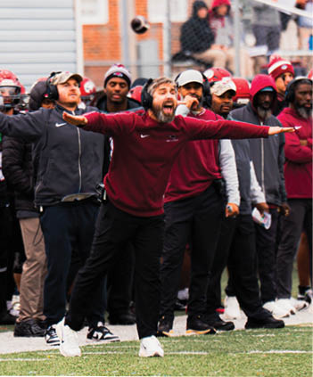 A person wearing a headset and maroon sweatshirt is energetically gesturing with arms outstretched while standing on a sports field with several other people in the background. Description generated by AI