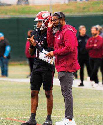 An adult wearing a red helmet and black sportswear stands next to another adult in a red jacket and gray pants holding a playbook and wearing a headset on a sports field. Description generated by AI