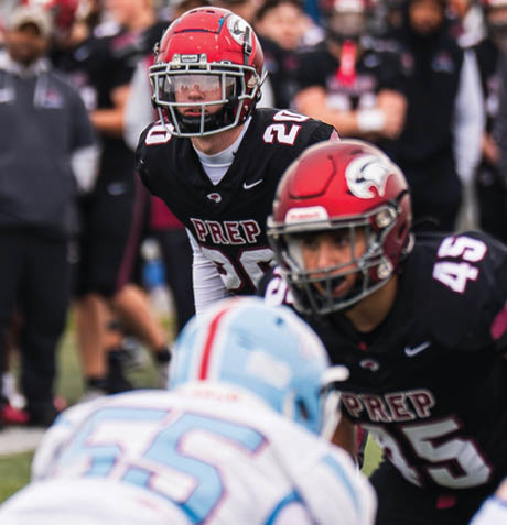 Two people in black football uniforms with red helmets are focused on a person in a white and blue football uniform during a game. Description generated by AI