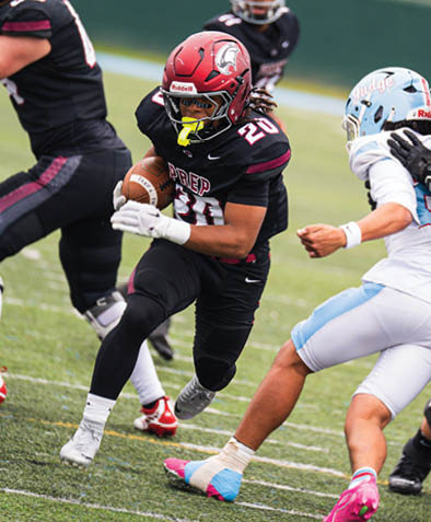 A person wearing a black football uniform with the number 20 is running with the ball while being pursued by a person in a white and light blue uniform on a grassy field. Description generated by AI