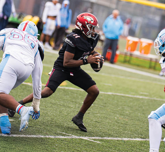 A person in a black football uniform is holding a football and preparing to throw while being approached by two people in white and light blue football uniforms on a grassy field. Description generated by AI