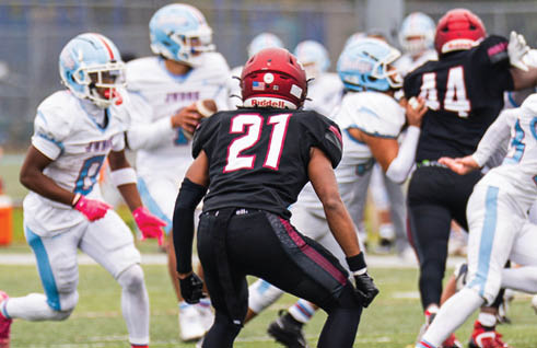 An adult wearing a black football uniform with the number 21 is crouched on a field facing several adults in white and light blue football uniforms during a game. Description generated by AI