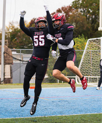 Two people in black athletic uniforms with red and white accents are jumping on a sports field with a blue and green surface near a soccer goal. Description generated by AI