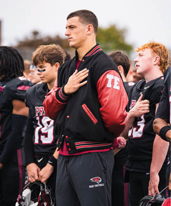 People wearing football uniforms stand with one person in a letterman jacket holding a hand over their chest, likely during a pre-game or national anthem moment. Description generated by AI