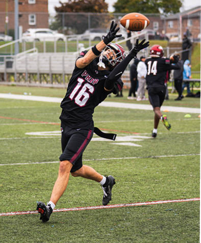 A person wearing a black football uniform with the number 16 is reaching up to catch a football on a grassy field while another person in a similar uniform stands in the background. Description generated by AI