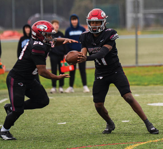 Two people in football uniforms and helmets are on a field with one handing off a football to the other while three people stand in the background near a fence. Description generated by AI
