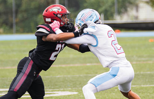 Two people in football uniforms are engaged in a blocking action on a grassy field with a fence and trees in the background. Description generated by AI