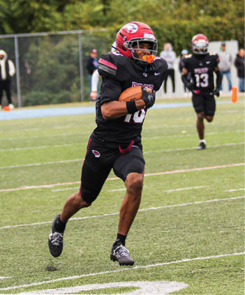 An adult wearing a black and red football uniform is running on a field while holding a football, with another player in the background. Description generated by AI