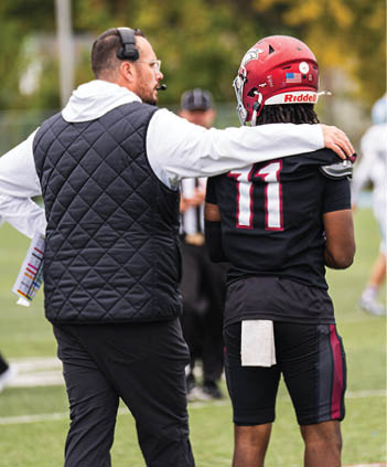 A person wearing a headset and a quilted vest has their arm around a person in a football uniform with the number 11 on the back, standing on a field. Description generated by AI