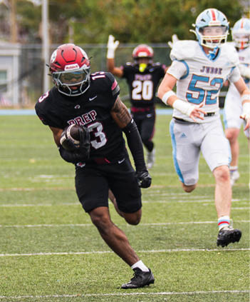 A person in a black football uniform with a red helmet is running on a field while holding a football, with two other players in the background. Description generated by AI