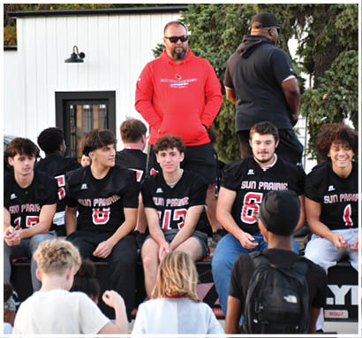 A group of people wearing football uniforms are on a field with one person in a white and green uniform standing in the center and others in red uniforms around them. Description generated by AI