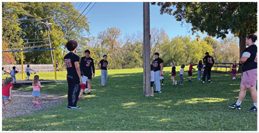 A group of people wearing football uniforms are on a field with one person in a white and green uniform standing in the center and others in red uniforms around them. Description generated by AI