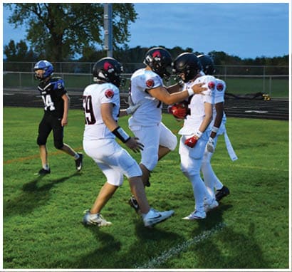 A group of people wearing football uniforms are on a field with one person in a white and green uniform standing in the center and others in red uniforms around them. Description generated by AI