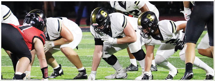 Several people wearing football helmets and uniforms are positioned on a field, appearing to prepare for a play during a nighttime game. Description generated by AI