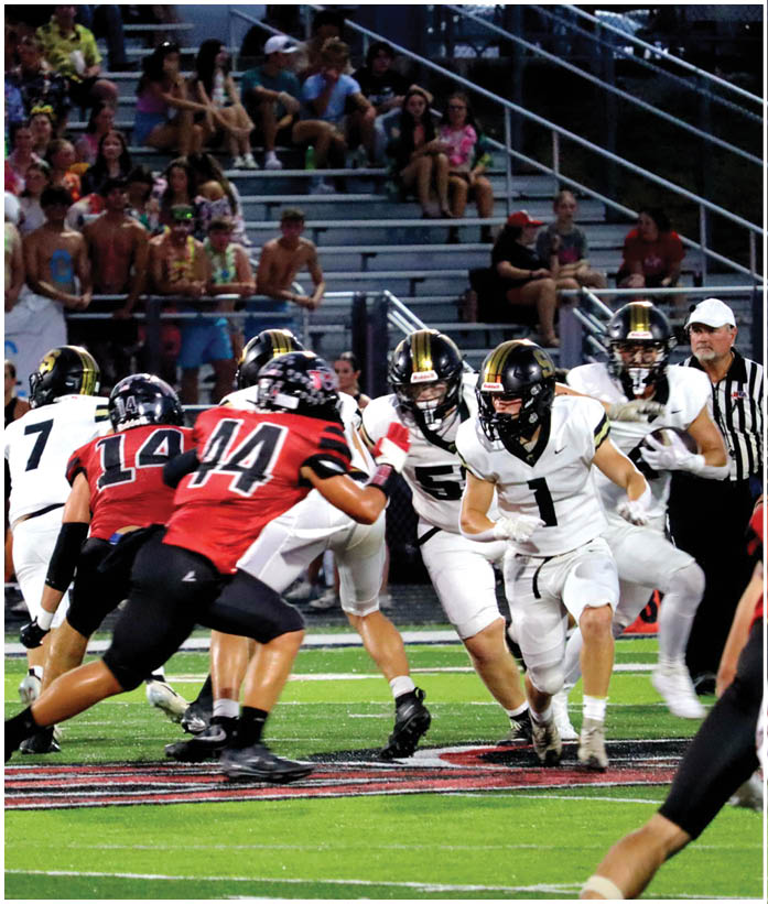 Two teams of adults in football uniforms are actively engaged in a play on a green field with spectators watching from bleachers in the background. Description generated by AI
