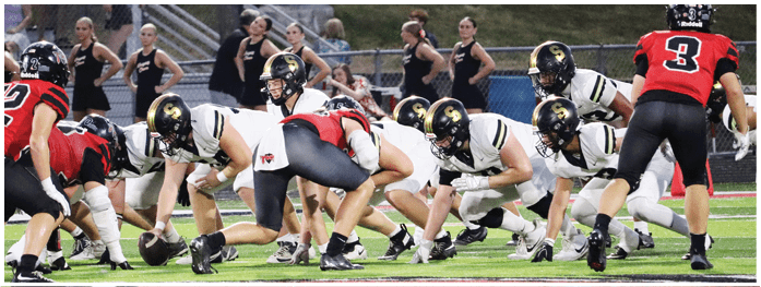 People in football uniforms are lined up on a field facing each other in a game formation with cheerleaders standing in the background. Description generated by AI