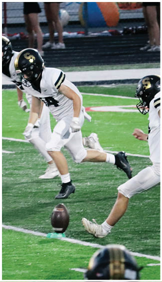 Three people in white football uniforms and black helmets are running on a green field as one of them kicks a football from a tee. Description generated by AI