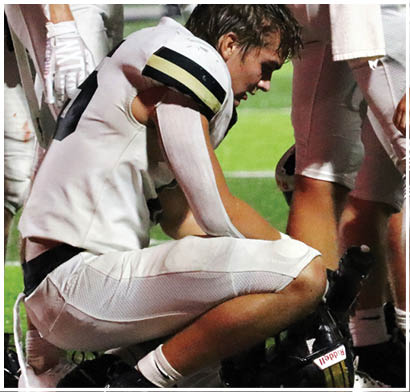 A person wearing a white football uniform with black and gold stripes is bent over on a field, appearing to be catching their breath or resting. Description generated by AI