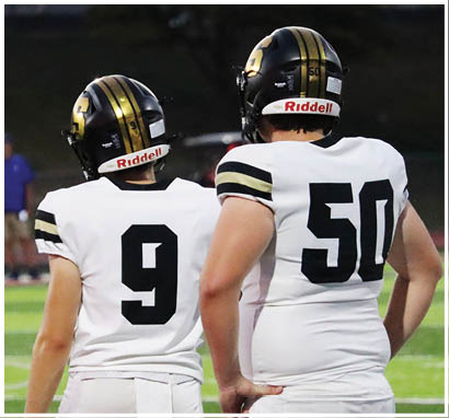 Two people wearing white football jerseys with black numbers and black helmets stand side by side on a field. Description generated by AI