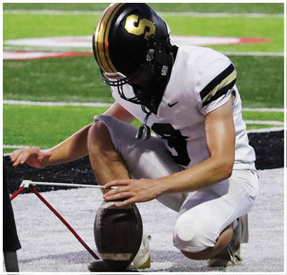 A person wearing a black helmet with gold stripes and a white sports jersey with black and gold accents is on a green field. Description generated by AI