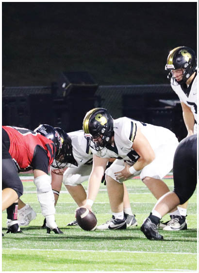 Two people in white football uniforms and helmets are positioned on a field, with one holding a football ready to snap and the other standing behind in a quarterback stance, while a third person in a red and black uniform is crouched in front of them. Description generated by AI