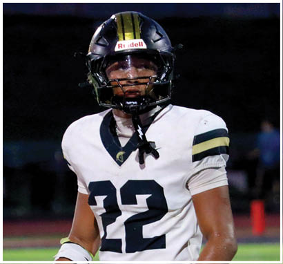 A person wearing a white football jersey with black and gold stripes and a black helmet with a gold stripe is standing on a field at night. Description generated by AI
