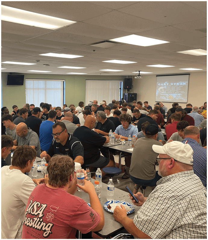 A large group of people seated at long tables in a cafeteria-style room are eating, drinking, and engaging in conversation. Description generated by AI