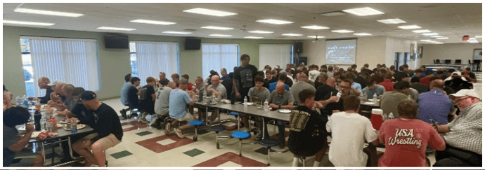 People sit at cafeteria tables with a tiled floor featuring a pattern of large red squares with green accents. Description generated by AI