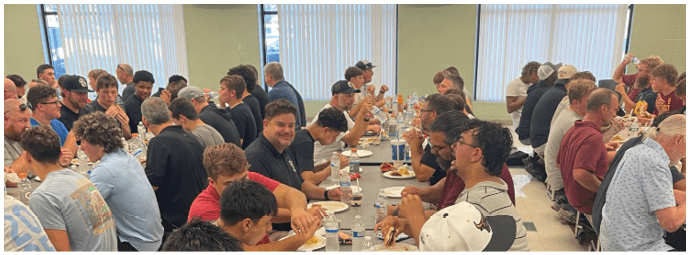 A group of people sitting closely together at a long table enjoying a meal with plates, drinks, and snacks in a brightly lit room. Description generated by AI