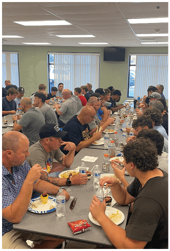 Two people sit at a table with plates of food, a water bottle, and a bag of Doritos chips between them. Description generated by AI