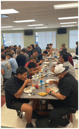 A large group of people sitting closely together at long tables in a cafeteria setting, eating and engaging in conversation. Description generated by AI