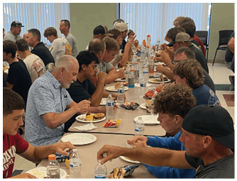 A large group of people sitting closely together at long cafeteria tables eating meals and drinking bottled water in a well-lit room with windows and televisions on the walls. Description generated by AI