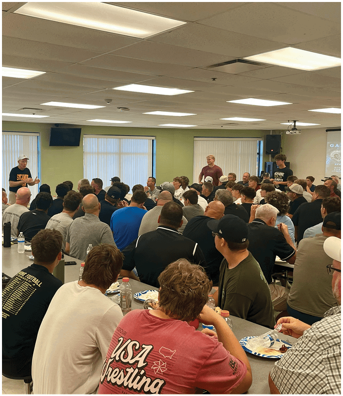 A large group of people seated at long tables in a cafeteria-style room are attentively listening to two people standing and speaking near the windows. Description generated by AI