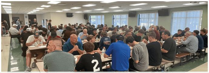 A large group of people seated closely together at long tables in a well-lit room, engaged in eating and conversation. Description generated by AI