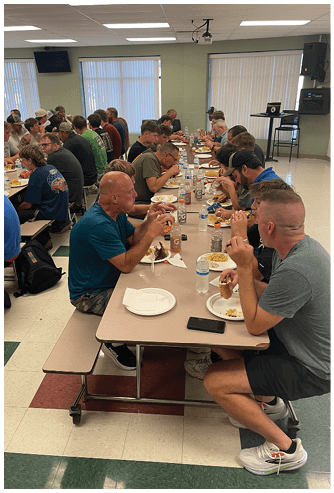 A large group of people sitting closely together at long cafeteria tables eating a meal in a brightly lit room. Description generated by AI