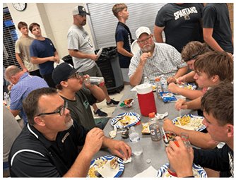 Several people sit around a table eating food from paper plates and drinking beverages in a casual indoor setting. Description generated by AI