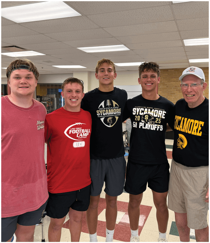 Five adults wearing casual clothing and sports-themed shirts stand closely together indoors on a checkered floor with tables and chairs in the background. Description generated by AI