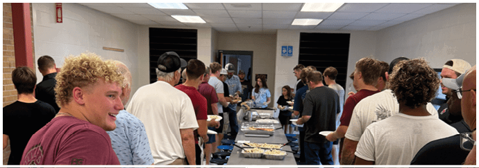 Adults are gathered around a long table with food trays in a room with tiled floors and a drop ceiling. Description generated by AI