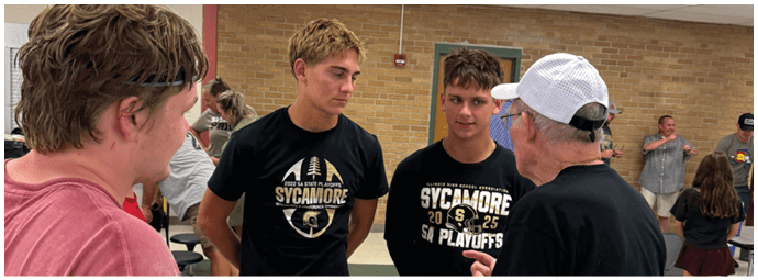 Four people are engaged in conversation in an indoor setting, with two wearing matching black shirts featuring a sports team logo and text about playoffs. Description generated by AI