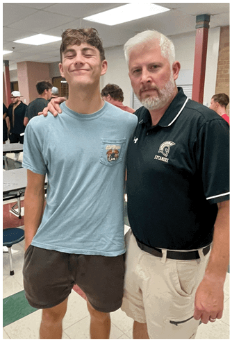 Two people stand closely together in a room with tables and chairs, one wearing a light blue t-shirt and shorts smiling with eyes closed, and the other wearing a black polo shirt with a logo and light-colored pants looking at the camera. Description generated by AI